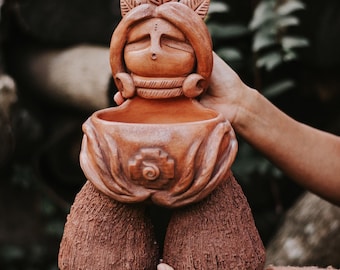 May include: Terracotta planter shaped like a woman with a crown of leaves, a detailed face, and a pot in her torso. The planter has a textured, rough surface and a spiral design on the front.
