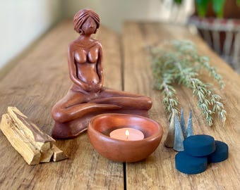 May include: A terracotta figurine of a woman in a seated meditation pose, holding her belly. The figurine is surrounded by a small terracotta bowl with a lit candle, palo santo sticks, incense cones, and dried herbs.