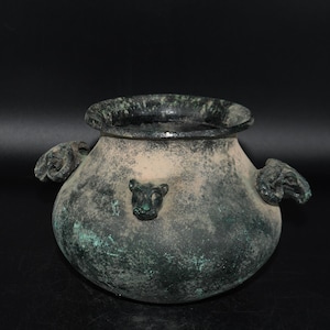May include: An antique bronze vessel with a wide, rounded body and a dark, textured rim. The vessel features two ornate handles and a small animal head detail. The bronze has a weathered, greenish patina, suggesting age and exposure to the elements.