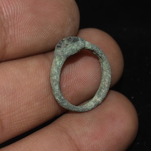 Puede incluir: Un pequeño anillo antiguo con una banda redondeada y un engaste en relieve. El anillo tiene una pátina marrón verdosa desgastada, que sugiere antigüedad. El engaste parece tener un elemento más oscuro, posiblemente una piedra.