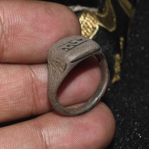 Puede incluir: Un anillo antiguo y desgastado, de color plateado apagado, con una cara rectangular. El anillo tiene grabados en la cara y los lados, que parecen ser una escritura o símbolo antiguo. El anillo está sostenido en una mano.