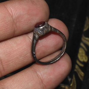 Puede incluir: Anillo de plata antiguo con una pequeña gema roja ovalada. El anillo tiene una banda detallada y texturizada y un engaste de bisel para la piedra. El anillo es probablemente vintage o antiguo.