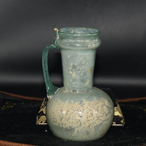 Puede incluir: Una jarra de vidrio verde claro vintage, con base redondeada y asa. La jarra tiene una superficie ligeramente texturizada con zonas descoloridas. El asa es de un tono verde más oscuro. Probablemente sea una antigüedad o una réplica.
