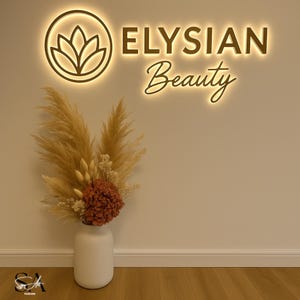 K&ouml;nnte beinhalten: Eine Wandtafel mit den Worten "ELYSIAN Beauty" in einer warm leuchtenden Schriftart. Darunter steht eine wei&szlig;e Vase mit einem Blumengesteck aus getrocknetem Pampasgras und einer rotbraunen Blume. Das Schild zeigt ein kreisf&ouml;rmiges Logo mit einer stilisierten Blume.