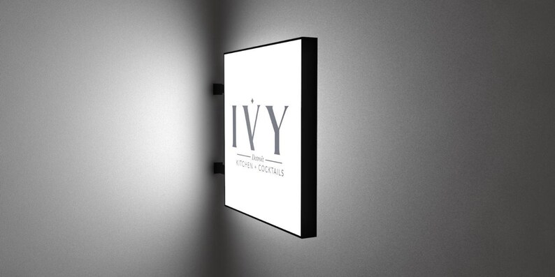 May include: A white rectangular sign with black trim and the text "IVY - Dining - Kitchen & Cocktails" on a white wall.