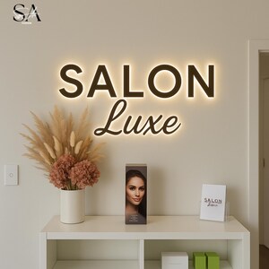 K&ouml;nnte beinhalten: Ein beleuchtetes Schild mit den Worten "SALON Luxe" in dunkler Schrift an der Wand. Darunter befindet sich ein wei&szlig;es Regal mit einer Vase mit Trockenblumen, einer Produktbox mit einem Frauengesicht und einem kleinen Schild. Das Ambiente ist modern und elegant.