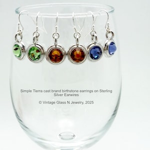 May include: Six gemstone earrings with silver earwires are displayed in a clear wine glass. The earrings feature round, colorful gemstones in green, orange, and blue hues. The text on the image says "Simple Tierra cast brand birthstone earrings on Sterling Silver Earwires."