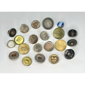 Puede incluir: Una colección de veintiún botones de metal variados en diferentes tamaños, formas y acabados. Varios botones presentan diseños en relieve, incluyendo locomotoras de vapor y los textos "Bartel", "Hercules" y "Thomas Co. Winner" grabados.
