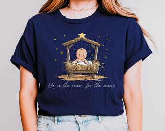 May include: Navy blue t-shirt featuring a cartoon illustration of a baby in a manger under a star. The text "He is the reason for the season" is written below the image. The shirt is a casual, everyday wear item.