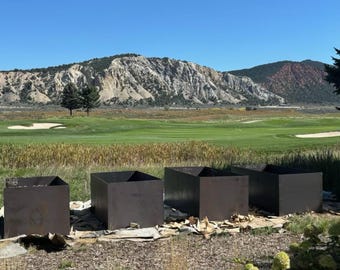 May include: Four black metal planters are arranged in a row on a grassy area in front of a golf course. The planters are empty and are set against a backdrop of a mountain range.