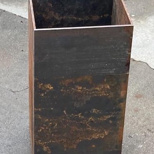 May include: A square, rusty metal planter with a dark brown finish. The planter is empty and sitting on a concrete surface.