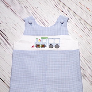 May include: A light blue and white gingham romper featuring a white chest panel with an embroidered train design. The train is light blue with green, yellow, and red accents. The romper has two fabric-covered buttons at the shoulder straps.