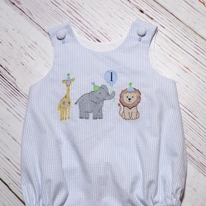 May include: A light blue and white gingham bubble romper featuring an embroidered design of a giraffe, an elephant holding a blue balloon with the number 1, and a lion, each wearing a small party hat, with two fabric-covered buttons at the shoulders.
