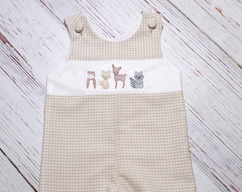 May include: Tan and white gingham baby romper with embroidered woodland animals. The romper features a white panel with embroidered fox, cat, deer, and raccoon designs. It has button closures on the straps and at the legs.