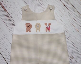 May include: A beige and white gingham baby romper with three embroidered dogs on the front. The dogs are brown, white, and black with a red bandana.