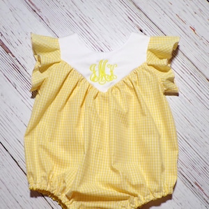 May include: Yellow and white gingham baby romper with a white bib-style neckline and ruffled sleeves. The romper features a yellow embroidered monogram. The garment is designed for infants and toddlers.