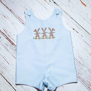 May include: Light blue baby overalls with three embroidered brown bunnies on the front. The overalls have a button closure at the shoulders and snaps at the bottom.