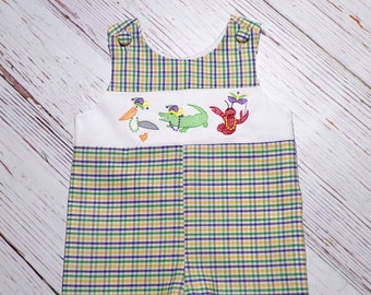 May include: A baby romper with a white bib featuring embroidered cartoon characters: a pelican, alligator, and crawfish, all wearing purple, green, and gold Mardi Gras hats. The romper has a plaid pattern in green, purple, and yellow.