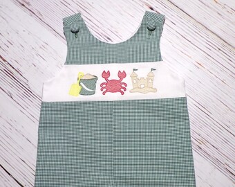 May include: Green and white gingham baby overalls with a white embroidered panel featuring a bucket, shovel, crab, and sandcastle.