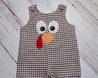 May include: Brown and white gingham overall romper with a turkey face applique. The turkey has red, orange, and white details.
