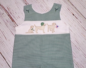 May include: Green and white gingham baby overalls with a white embroidered panel featuring three dogs. The dog in the middle is wearing a military helmet and the dog on the right is holding a small American flag.