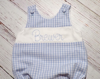 May include: A blue and white gingham baby onesie with a white band across the chest and the name "Brewer" embroidered in cursive.