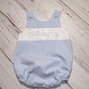 May include: A blue and white gingham baby onesie with the words "Birthday Boy" embroidered on the front.