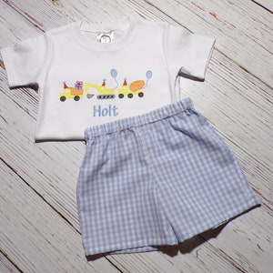 May include: A white short-sleeved shirt with a yellow construction truck design and the name "Holt" embroidered on the front. The shirt is paired with blue and white gingham shorts.