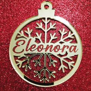 Puede incluir: Adorno navideño de color dorado con un diseño de copo de nieve y el nombre "Eleonora" escrito en él.