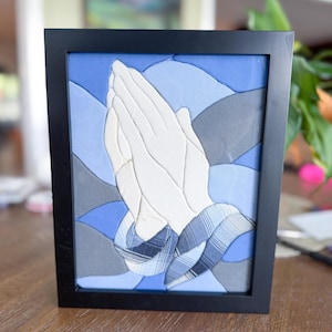 May include: A framed wall art piece featuring a fabric collage of praying hands. The hands are made of off-white fabric, while the background consists of layered blue and grey fabric shapes. The cuffs are made of a blue and white plaid fabric.