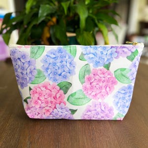 Puede incluir: Un neceser rectangular con un estampado floral de hortensias rosas, moradas y azules y hojas verdes sobre un fondo blanco. Tiene un cierre de cremallera dorado.