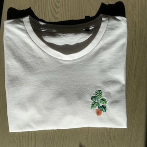 May include: White t-shirt with a small embroidered green plant in a brown pot.