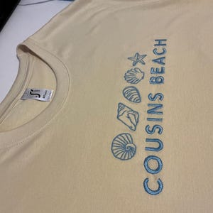 Peut inclure: Un t-shirt beige clair à col rond. Le t-shirt présente le texte "COUSINS BEACH" en bleu, ainsi que des motifs brodés de coquillages et d'étoiles de mer bleus. Le t-shirt est fait d'une matière douce.