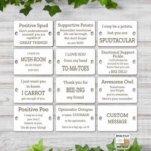 May include: A set of 12 white wooden tags with various positive messages and puns. Each tag has a hole at the top for hanging. The tags are arranged in a grid pattern on a light wood surface.