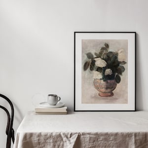 Neutral colors Painting of a bouquet of white roses, framed and displayed on a table covered with a linen tablecloth, a chair and a cup of coffee on top of a pile of book. | Dear Alice Vintage Art