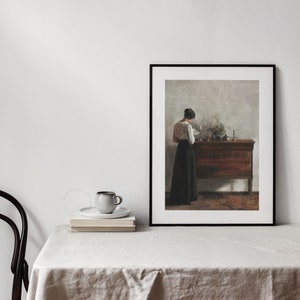 Woman Reading Painting framed and displayed on a table with a linen tablecloth, a black chair and a pile of book | Dear Alice Vintage Art