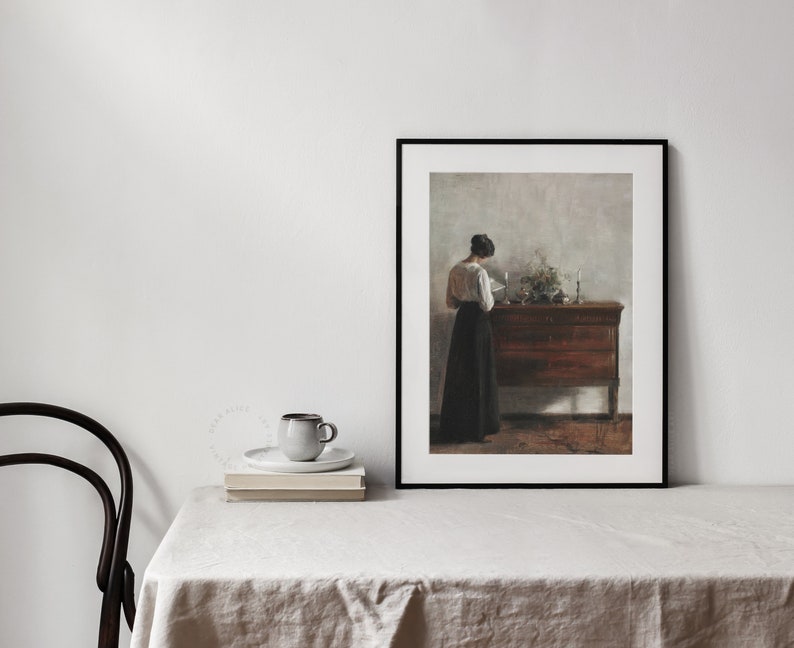 Woman Reading Painting framed and displayed on a table with a linen tablecloth, a black chair and a pile of book | Dear Alice Vintage Art