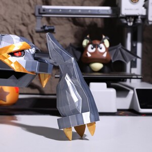 May include: A 3D printed model of a gray and gold robotic hand with sharp claws. The hand is posed as if it is reaching out. The model is in focus, while a 3D printer and a brown and black 3D printed model of a character are out of focus in the background.