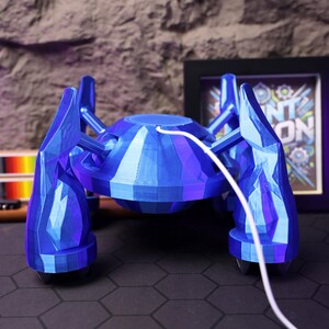 May include: A blue, geometrically-shaped, 3D printed robot with a white cord attached to the back. The robot has four legs and a round body. The robot is sitting on a black hexagonal tile floor.