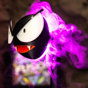 May include: A black and white 3D-printed figure of a Gastly, a Pok&eacute;mon character, with a red mouth and sharp teeth. The figure is surrounded by a cloud of purple smoke, creating a spooky effect.