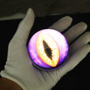 The LED crystal ball with custom magic eye is placed beside anime figures and gaming accessories, emphasizing its value as a holiday or birthday gift for him. Its crystal-clear orb and fiery visual inside enhance any gaming station, bookshelf, or nightstand, adding a touch of the supernatural with its gothic design and fantasy-themed projection that’s perfect for cosplay or room decoration.