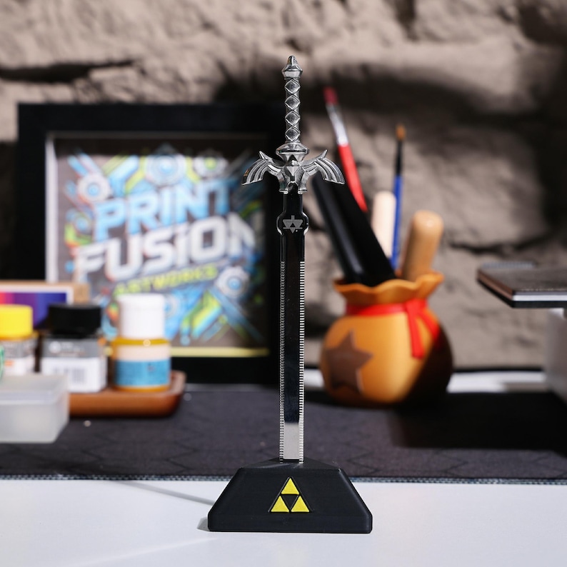 May include: A silver Master Sword replica with a black base featuring the Triforce symbol. The sword is on a table with a painting in the background.