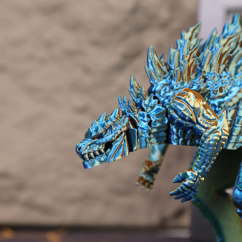 Angled top-down shot of the blue-green electroplated Godzilla figure displayed on a wooden shelf, surrounded by other collectible items. The photo captures the overall proportions of the figure, its vibrant metallic finish, and the attention to detail in the dorsal plates and body texture. This Godzilla figure is an excellent addition to any monster-themed room, gaming station, or collector display setup, designed to appeal to fans of Japanese kaiju, sci-fi films, and 3D printed art.