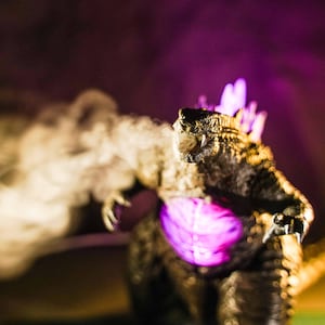 May include: A dark, textured Godzilla figure with a glowing purple chest and dorsal fins. The creature has its mouth open, emitting a thick, light-coloured cloud of smoke or vapour that extends horizontally across the frame to the left side.