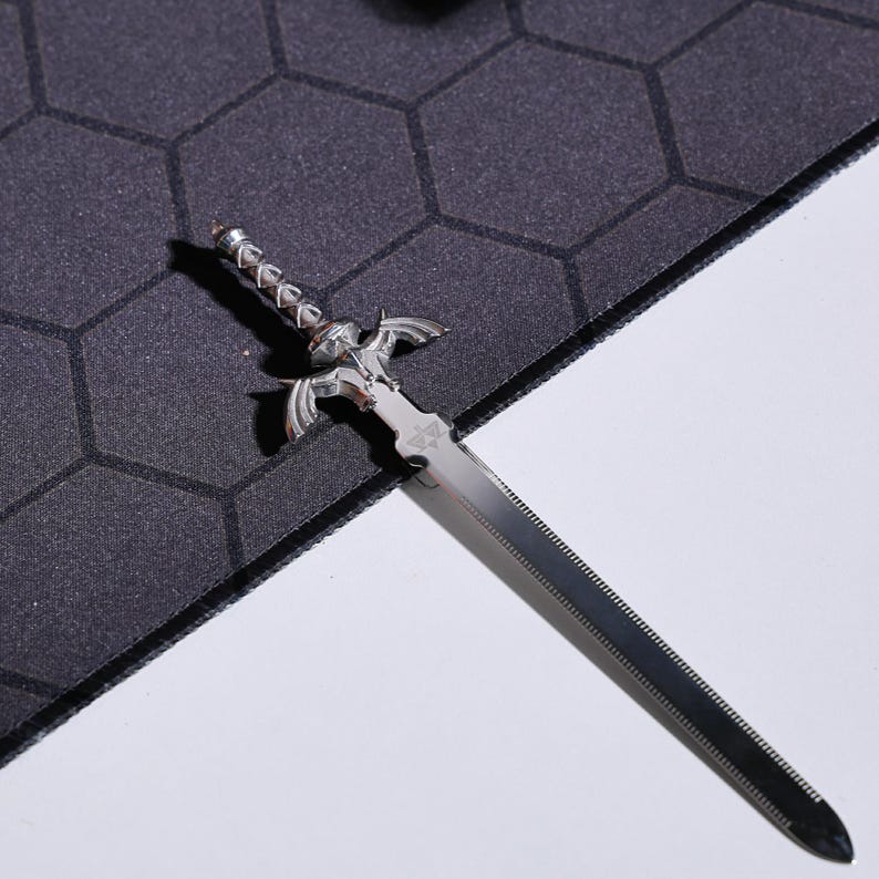 May include: A silver-colored sword with a detailed hilt and a serrated blade. The sword is lying on a black and gray textured surface.