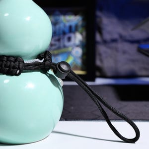 May include: A light blue ceramic gourd with a black paracord strap and a black plastic buckle. The strap is looped and tied around the gourd.