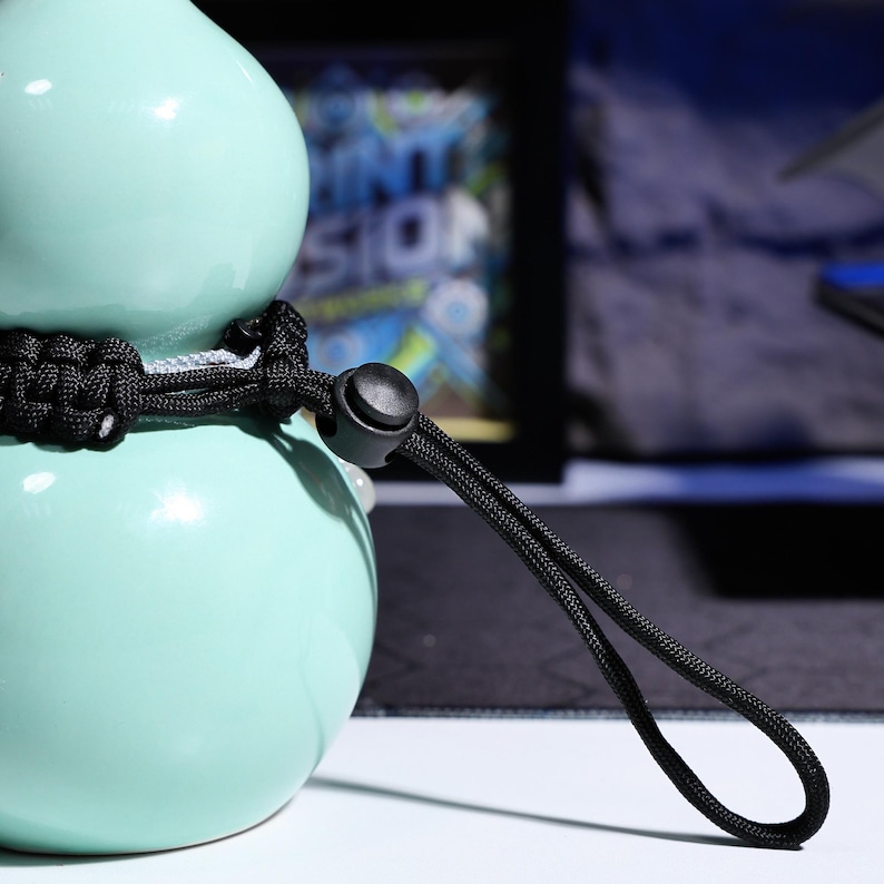May include: A light blue ceramic gourd with a black paracord strap and a black plastic buckle. The strap is looped and tied around the gourd.