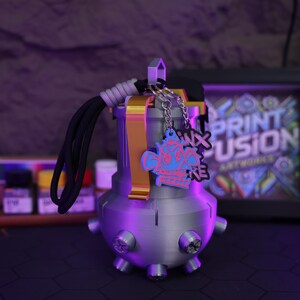 May include: A 3D printed gray grenade-shaped keychain with a gold accent and a black cord. The keychain has two charms attached to it, one with a pink and blue skull design and the other with the text "3D Print Fusion" in pink and blue.