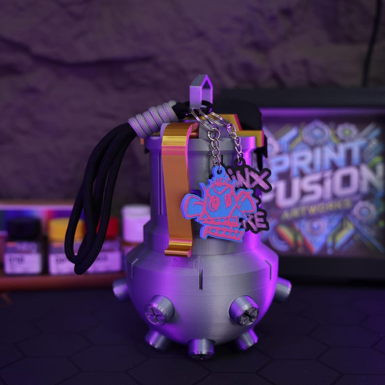 May include: A 3D printed gray grenade-shaped keychain with a gold accent and a black cord. The keychain has two charms attached to it, one with a pink and blue skull design and the other with the text "3D Print Fusion" in pink and blue.