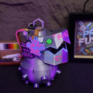 May include: A 3D printed gray grenade with a pink and blue heart and the word "BONJOUR" written on it. The grenade has a silver chain with a pink and blue charm that says "I WAS HERE".